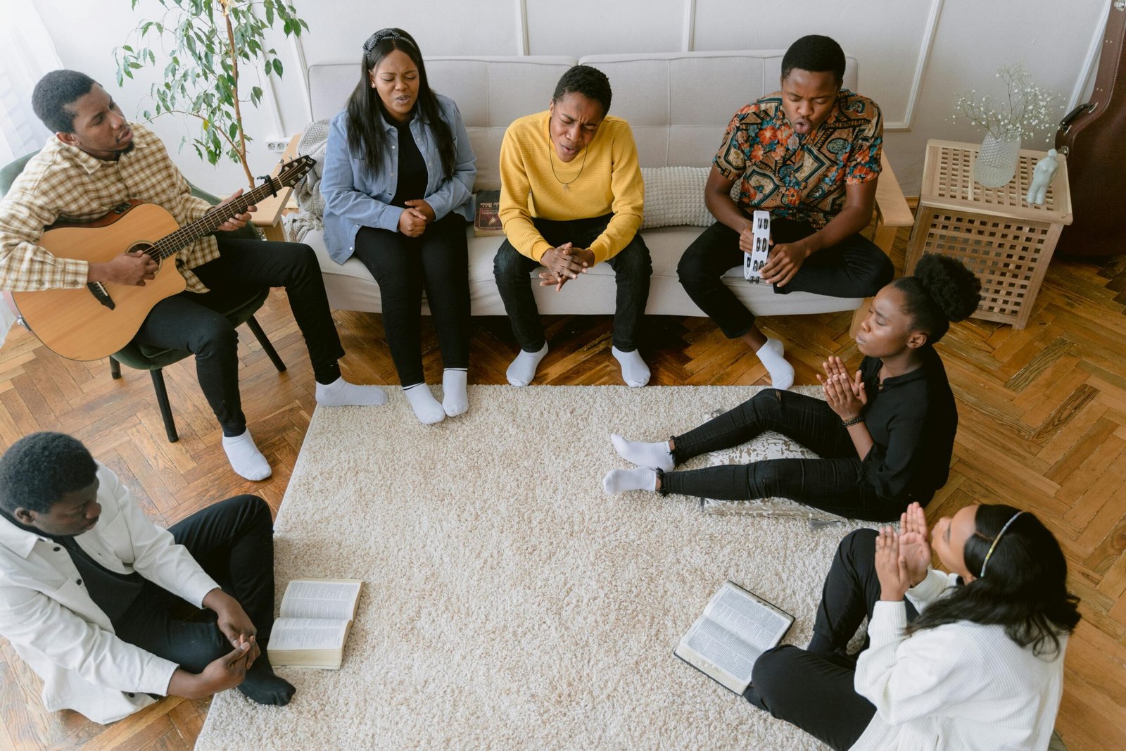 A diverse group of people praying, singing, and playing guitar indoors, emphasizing faith and community.